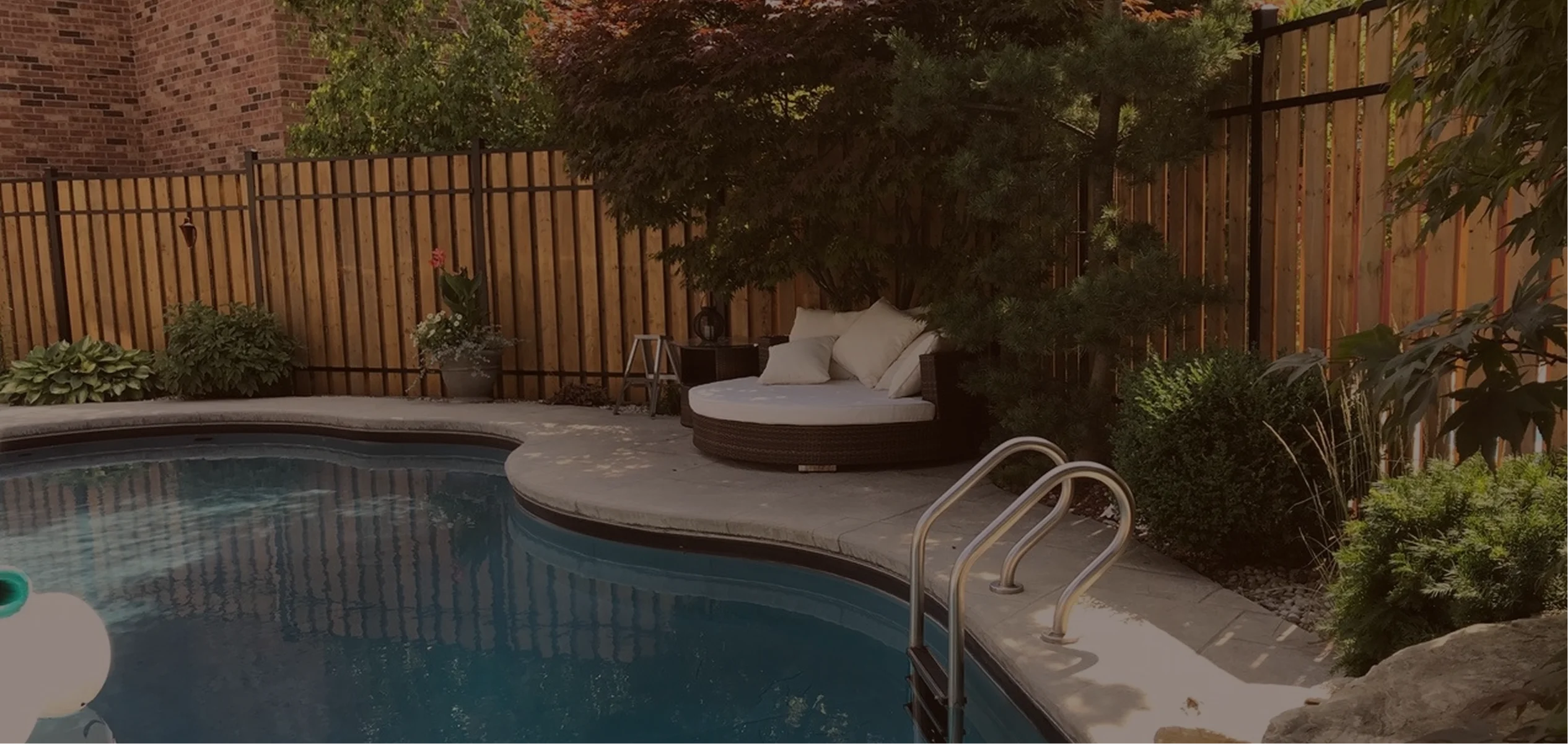 Backyard pool area with curved edge, a metal pool ladder, surrounded by shrubs, a wooden fence by slipfence, and a cushioned round wicker daybed with pillows under a tree.