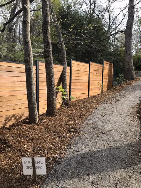 A gravel pathway runs alongside a fence by slipfence with trees growing close to it in a wooded area.