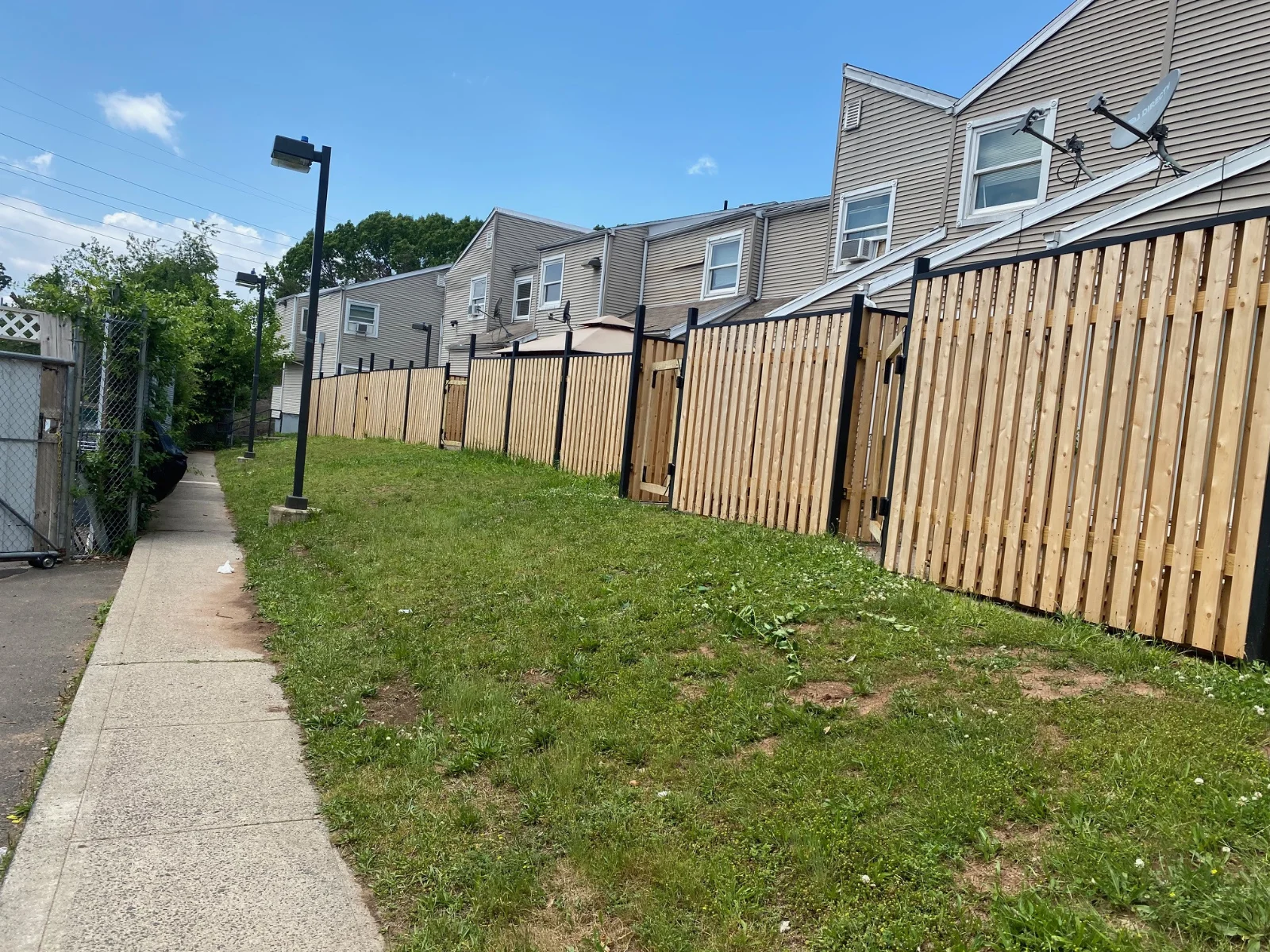 Sidewalk next to a grassy area with fences by Slipfence and townhouses under a clear blue sky.