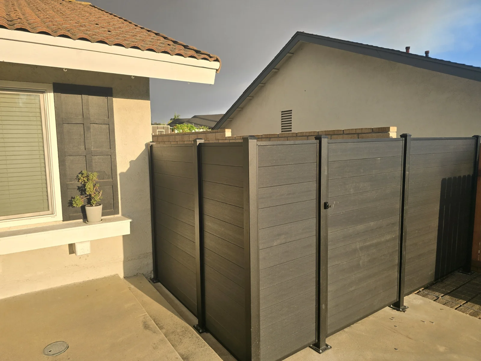 Outdoor corner of a house with a small potted plant on the windowsill and a dark privacy fence by Slipfence enclosing a concrete patio area.
