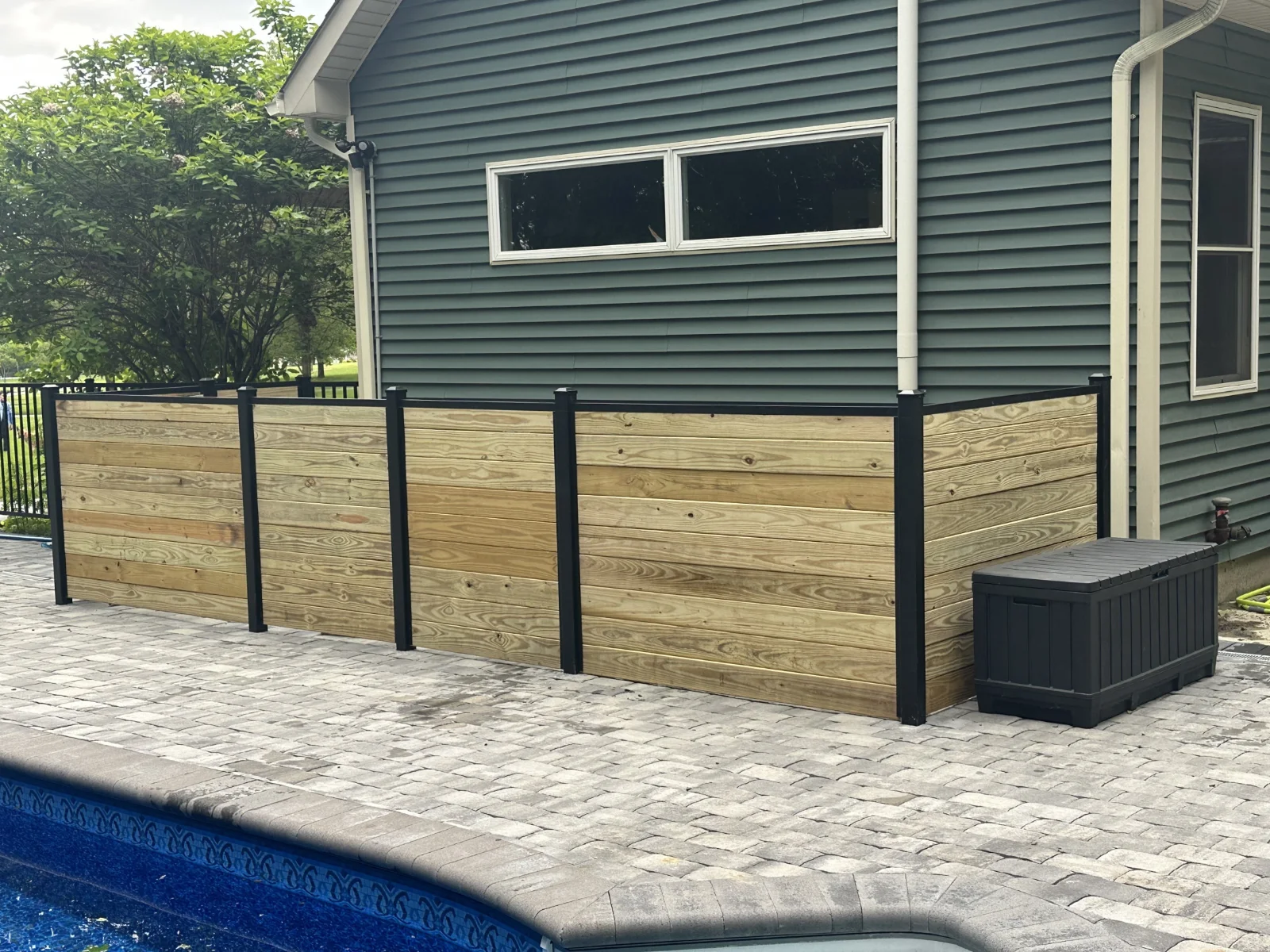 Outdoor poolside area with a stone tile deck, a privacy fence by Slipfence with black metal posts around a house corner, and a black storage box.