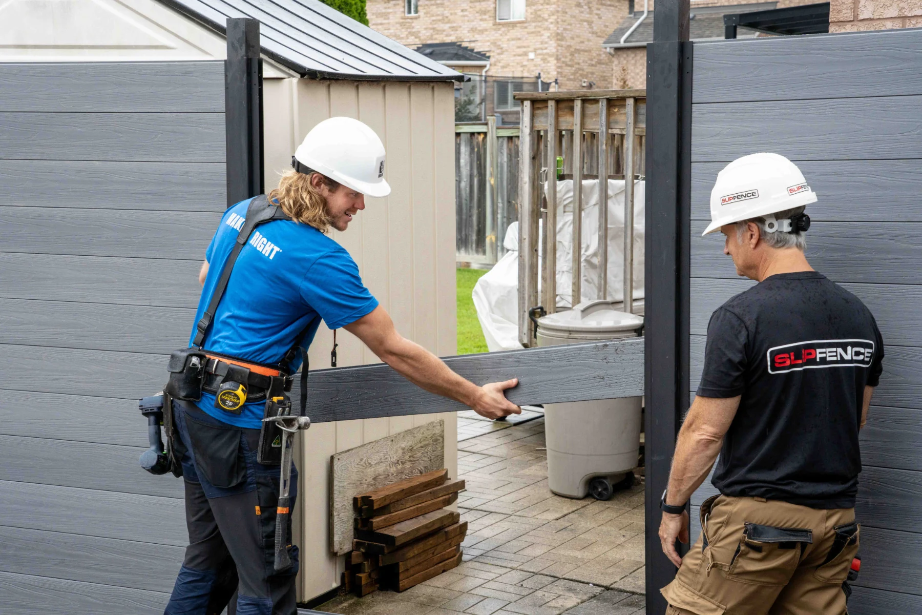 Two construction workers wearing hard hats installing a gray  fence panel by Slipfence outside in a residential backyard.