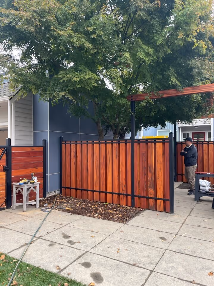 Person standing near a newly installed fence by Slipfence with black metal framing under a large leafy tree beside a paved sidewalk.