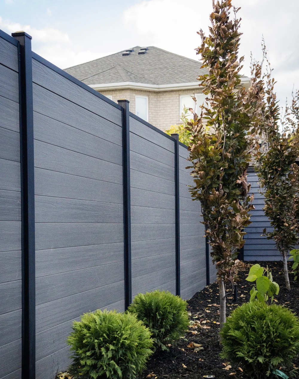 Modern gray wood-panel fence by Slipfence with black posts alongside a garden bed with shrubs and small trees.
