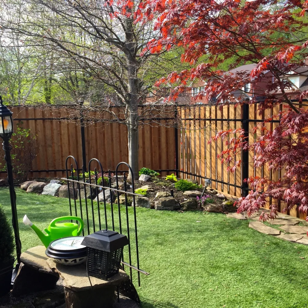 Backyard with green artificial grass, a stone bench holding a green watering can, clock, and black lantern, surrounded by a wooden fence by Slipfence and trees including one with red leaves.