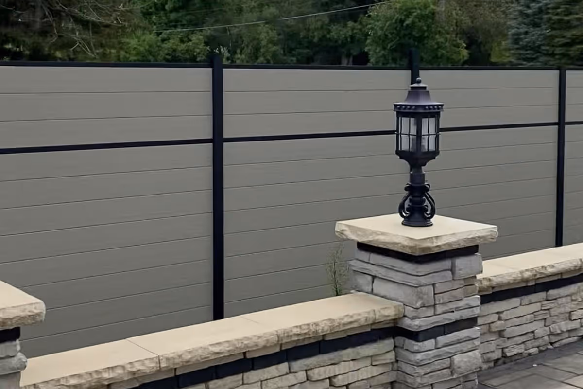 Stone patio wall by Slipfence with beige capstones and an ornate black lantern on a pillar in front of a gray privacy fence with black horizontal and vertical supports.