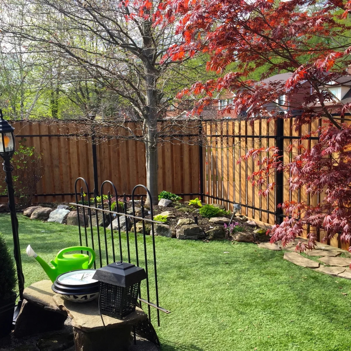 Backyard garden with green grass, a leafless tree, red-leafed tree, stone-bordered flower bed, and a green watering can on a stone table enclosed by a fence by Slipfence.