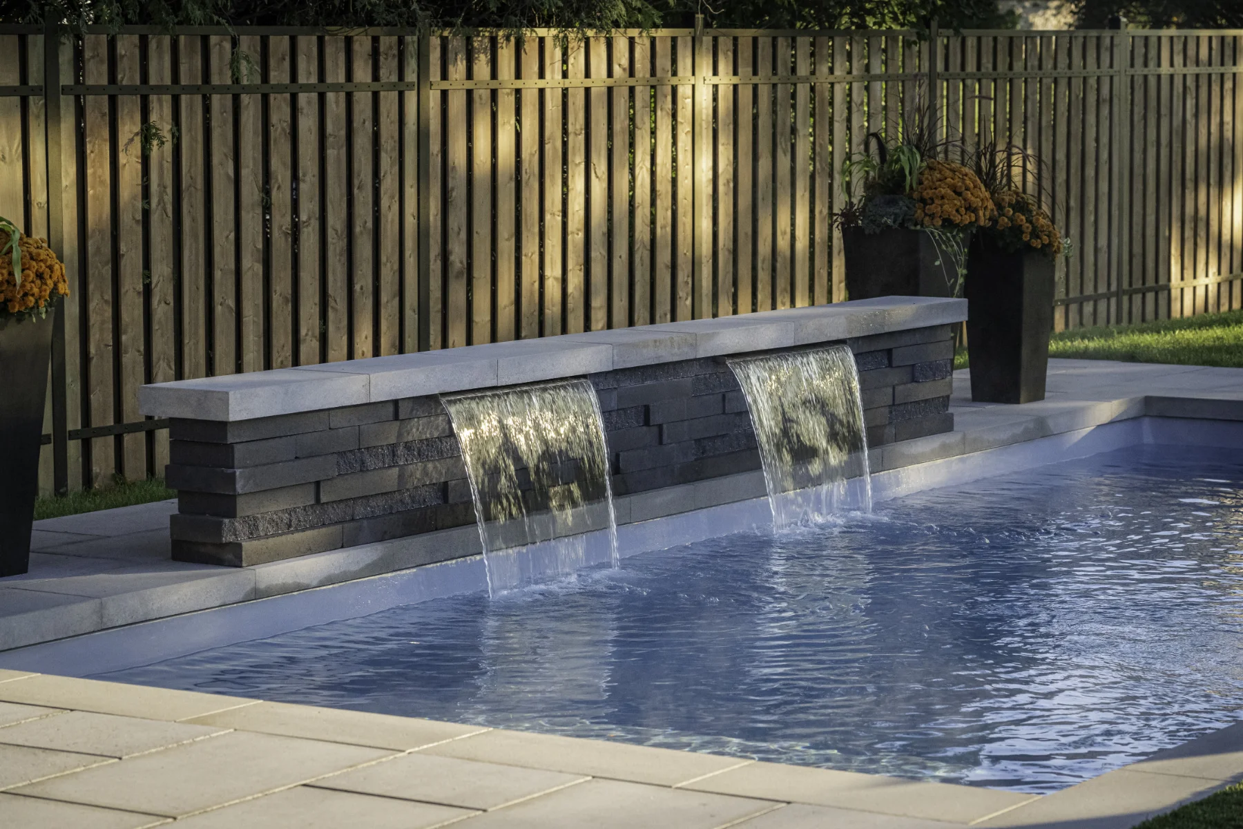 Swimming pool with two stone water fountains flowing water into the pool, bordered by a fence by Slipfence and flower planters.