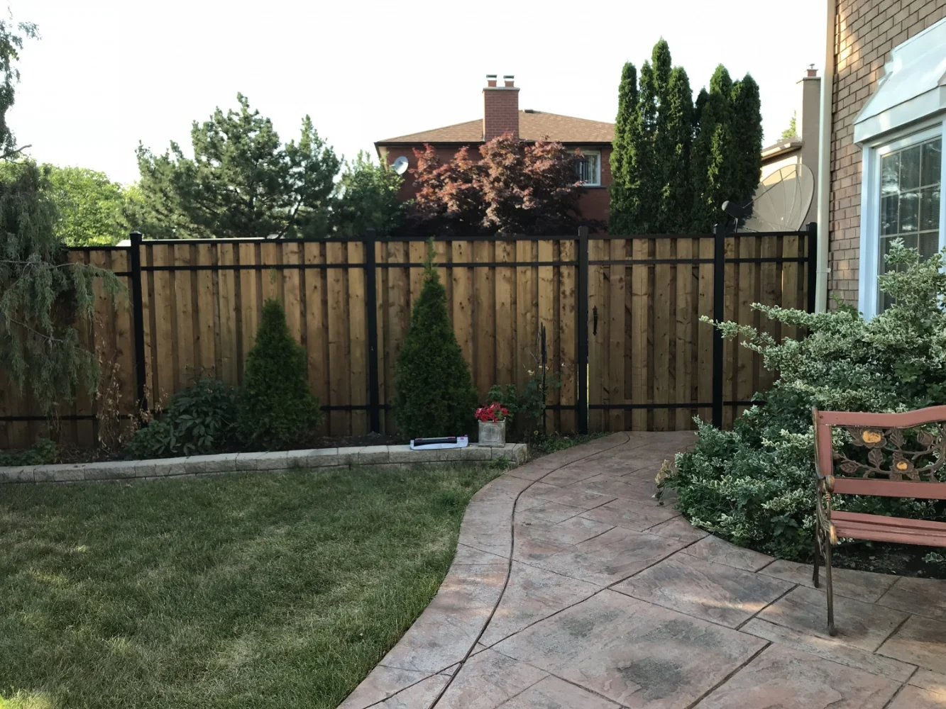 Backyard with a stone-paved walkway, fence by Slipfence, green grass, shrubs, and a decorative bench near a brick house.