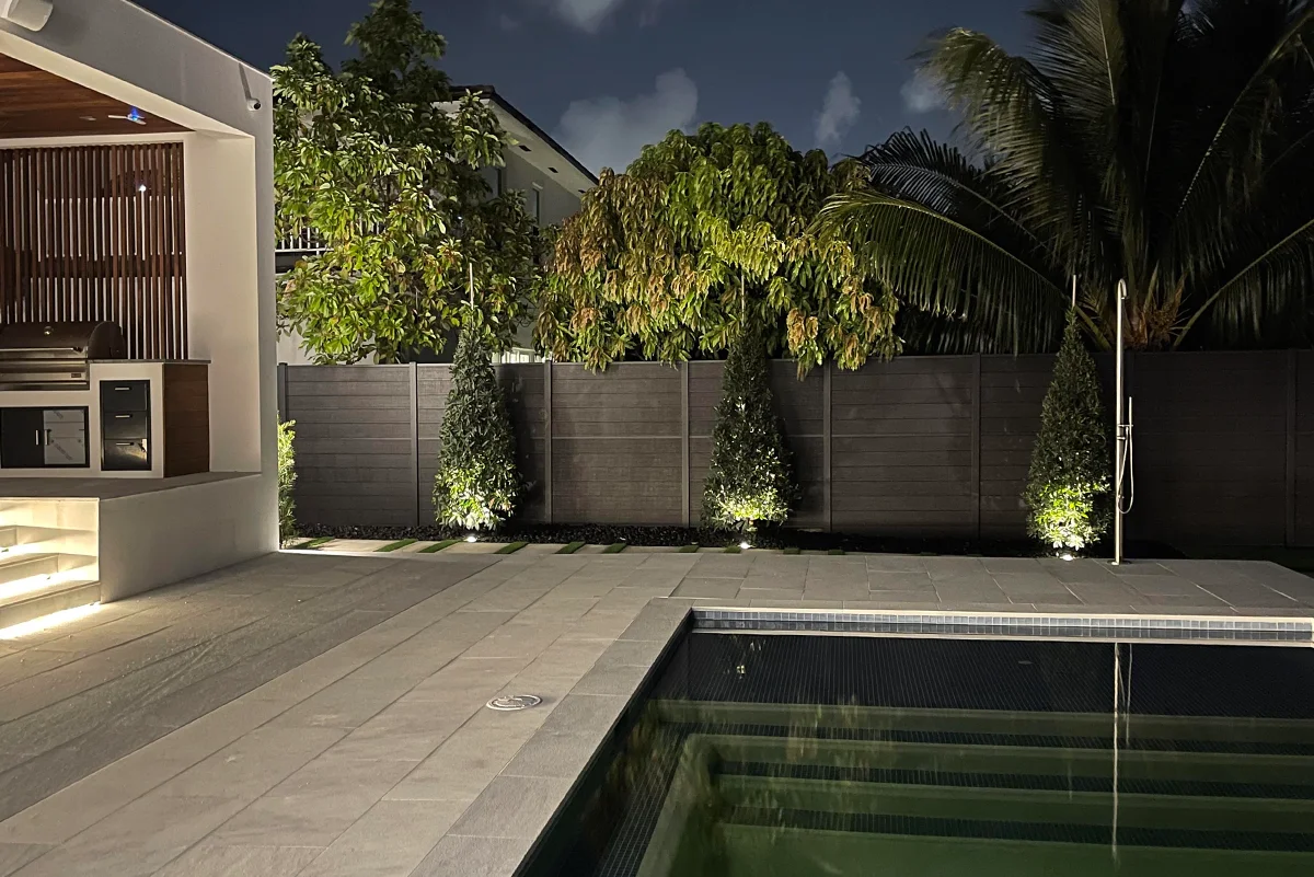 Night view of a modern outdoor patio with a lit swimming pool, tiled flooring, garden trees, and an outdoor kitchen area enclosed with a fence by Slipfence.