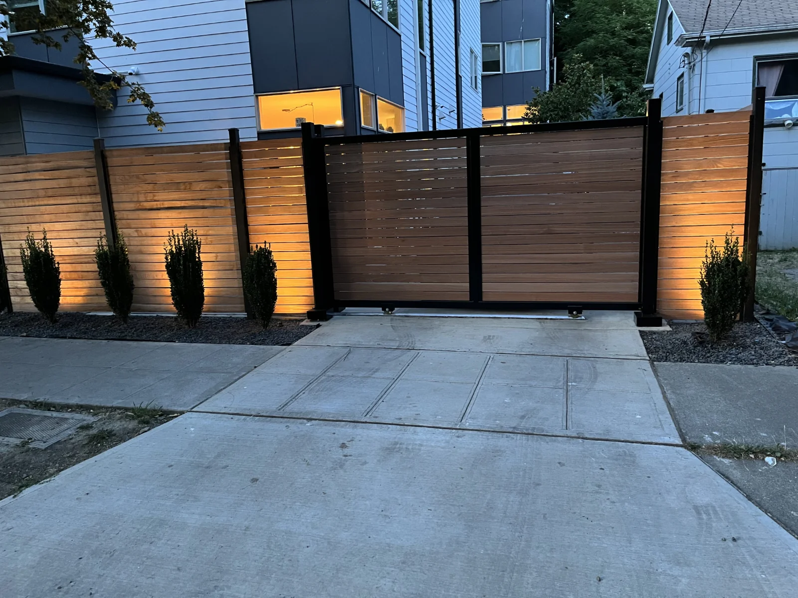 Wooden privacy fence by Slipfence and gate with black metal frame, illuminated at dusk with small bushes along the fence line.