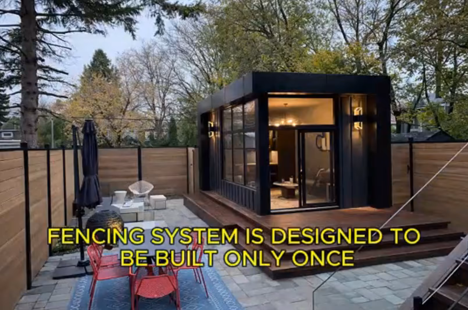 Modern backyard patio by Slipfence with black framed glass building, red chairs around a table on a blue rug, and wooden fencing.
